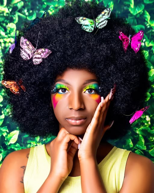 Woman with colorful makeup and butterflies in her afro against green foliage.