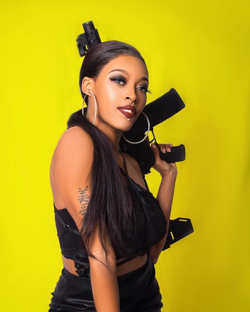 Woman in black with hoop earrings posing against a bright yellow studio background.