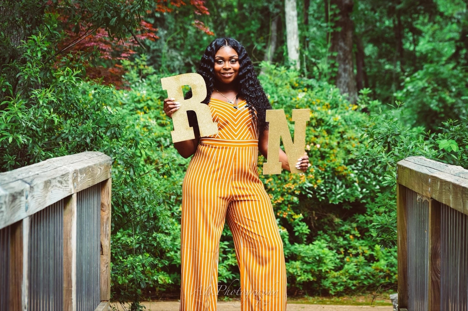 Nursing graduate in a yellow jumpsuit holding R and N letters in a garden.