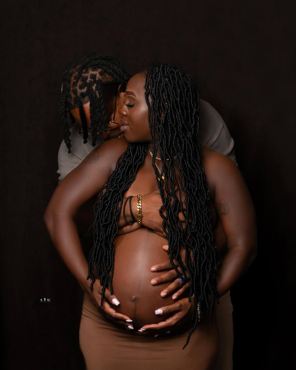 Couple embracing with their hands on her pregnant belly against a dark background.